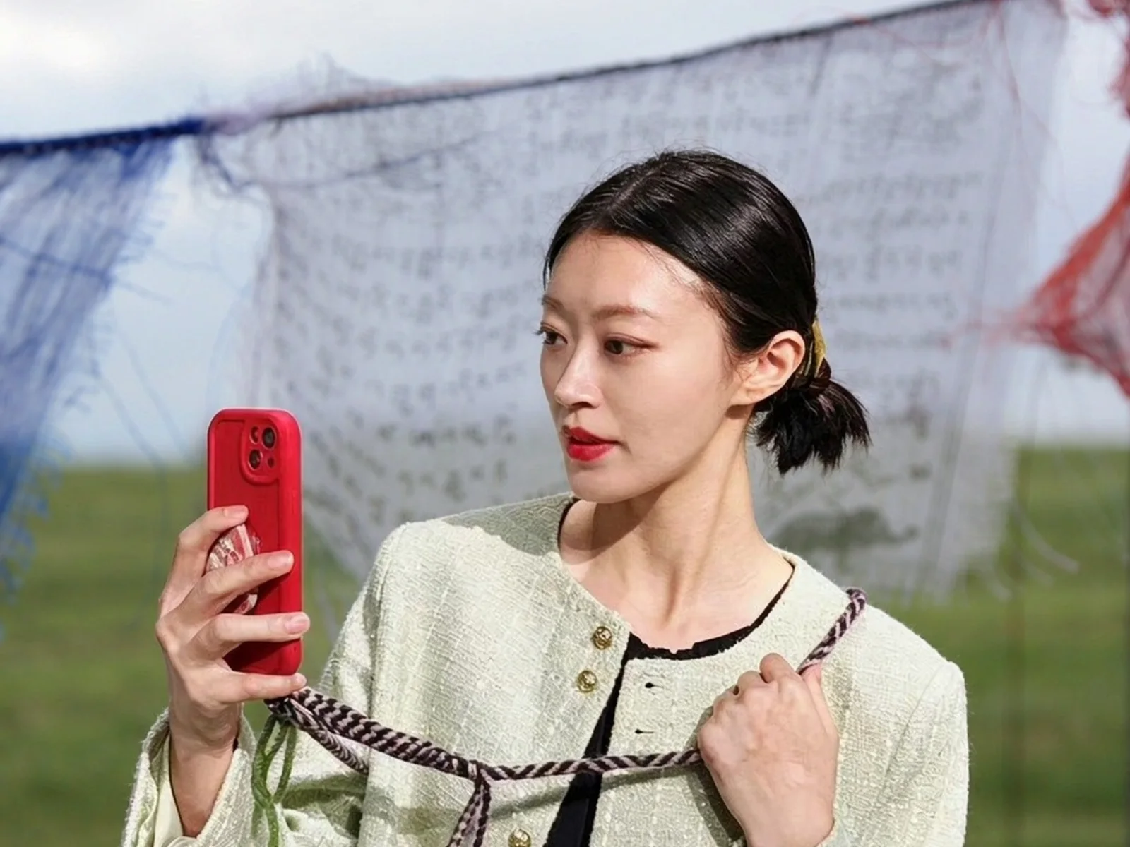 Nelly ZHUANG wearing a Dopamine wool wurdoo chain across the body, photographed on the Tibetan highland beneath a line of prayer flags