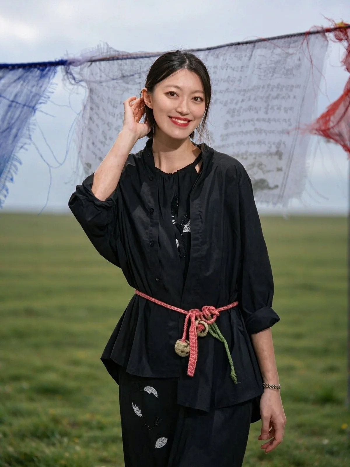 Nelly ZHUANG in a black top wearing a wurdoo chain as a waist wrap, prayer flags behind her on the highland